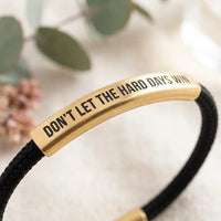 Macro detail of Don't Let The Hard Days Win 
gold engraved plate with black uppercase 
lettering on stainless steel — premium 
motivational bracelet close-up