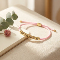 Don't Let The Hard Days Win bracelet in pink 
nylon cord with gold engraved plate on cream 
linen with eucalyptus and dried rose — 
motivational mental wellness bracelet